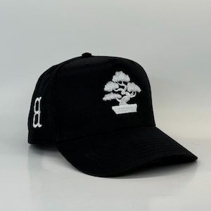 Puede incluir: Gorra de béisbol negra con un diseño de bonsái bordado en blanco en la parte delantera. La gorra también presenta una letra "B" blanca en el lateral. Tiene una visera curva y un botón en la parte superior.