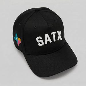 Può includere: Un cappellino da baseball nero con il testo SATX ricamato in bianco sul pannello frontale. Il lato del cappellino presenta una piccola toppa multicolore a forma dello stato del Texas, con sezioni blu, bianche, rosa e gialle.