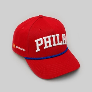 Peut inclure: Une casquette de baseball rouge vif avec le texte PHILA brodé en blanc sur le devant. Une bordure en cordelette tressée bleue se trouve à la base de la calotte. Le côté de la casquette affiche un carré orange avec la lettre B et le texte NRG Station.