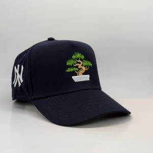 Könnte beinhalten: Eine marineblaue Baseballkappe mit einem weißen gestickten 'NY'-Logo auf der Vorderseite und einem grünen gestickten Bonsai-Baum auf der Krone.
