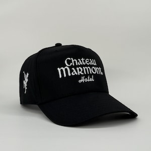 May include: Black baseball cap with the text "Chateau Marmont Hotel" embroidered in white. A white graphic is on the side of the cap. The cap has a curved brim and a button on top.