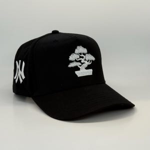 May include: A black baseball cap featuring a white embroidered bonsai tree design and a white embroidered "NY" logo on the side.