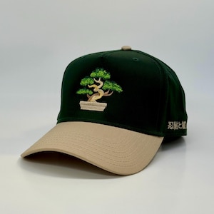 May include: A baseball cap with a dark green crown and a tan brim. The cap features an embroidered bonsai tree design in green and brown on the front. The text "忍耐と観" is embroidered on the side.