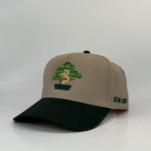 May include: Tan baseball cap with a dark green brim. The cap features an embroidered bonsai tree design in green and brown. Japanese characters are printed on the side of the cap.
