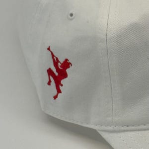 May include: White baseball cap with a red embroidered figure on the side. The cap is made of a smooth fabric and features a curved brim. The red figure appears to be a stylized martial arts pose.
