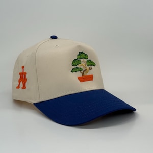 Könnte beinhalten: Baseballcap mit cremefarbener Krone und blauem Schirm. Die Kappe hat ein gesticktes Bonsai-Baum-Design in Grün, Braun und Orange. Auf der Seite befindet sich ein orangefarbenes Emblem.