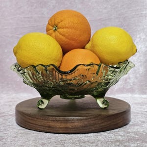 May include: A green glass fruit bowl with a decorative floral design, holding two oranges and two lemons. The bowl sits on a round, dark wooden base. The fruit bowl is a decorative item.