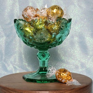 May include: A green glass candy dish filled with gold and yellow foil-wrapped chocolates. The dish has a decorative, cut-glass design and sits on a wooden surface. One chocolate rests beside the dish. The background is a light blue with glitter.
