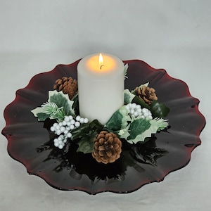 May include: A lit white pillar candle sits on a dark red, scalloped-edge glass plate, surrounded by pinecones, white berries, and green and white leaves. A festive centerpiece for the holidays.