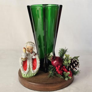 May include: A green glass vase with a dark base, a figurine of a girl holding a candle, and a decorative arrangement of a pomegranate, berries, pine needles, and a pine cone on a wooden base. The vase is tall and has a geometric design.