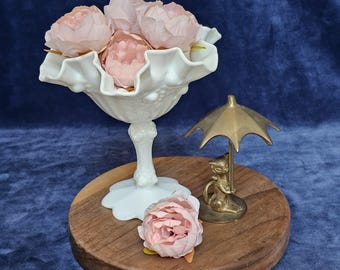 May include: A white, ruffled-edge glass bowl filled with pink flowers sits on a wooden tray. A small brass figurine of a cat holding an umbrella is next to the bowl. A single pink flower rests on the tray.