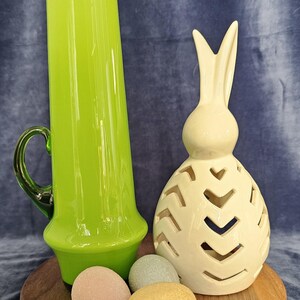 May include: A green glass vase with a handle sits on a wooden round base with three pastel colored Easter eggs. A white ceramic bunny with cut-out designs is next to the vase.