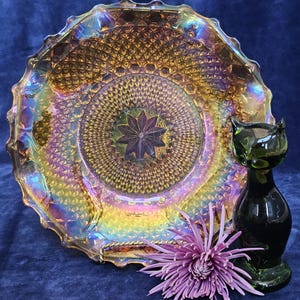 May include: An iridescent glass plate with a starburst design in the center, a green glass cat figurine, and a purple chrysanthemum flower. The plate has a textured surface and a scalloped edge. The cat figurine is sleek and modern.