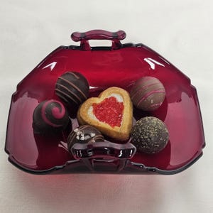 May include: A ruby red glass dish shaped like a purse, filled with assorted chocolates and a heart-shaped cookie. The chocolates are dark and milk chocolate, some with decorative swirls and stripes. The dish has a handle and a glossy finish.