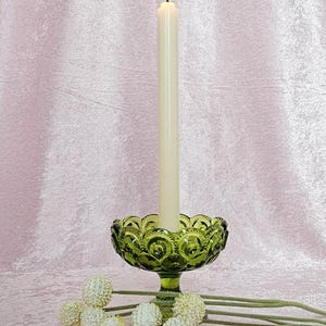 May include: A green glass candle holder with a pedestal base and a white candle burning inside. The candle holder is decorated with a pattern of small circles. The candle holder is sitting on a pink surface with white flowers.