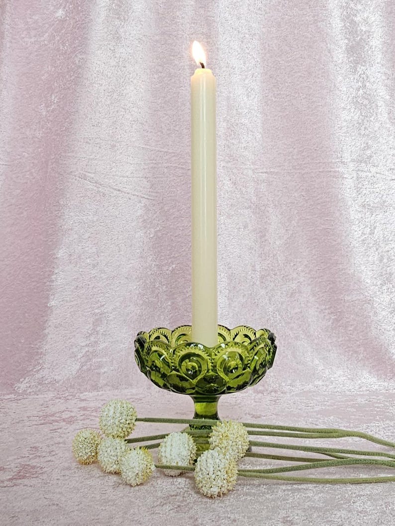 May include: A green glass candle holder with a pedestal base and a white candle burning inside. The candle holder is decorated with a pattern of small circles. The candle holder is sitting on a pink surface with white flowers.