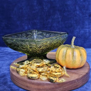 May include: A decorative arrangement featuring a green glass candy dish with floral patterns, a small orange pumpkin, and Werther's Original candies on a wooden tray. The background is a dark blue fabric.
