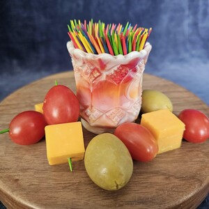May include: A decorative, opalescent glass toothpick holder filled with colorful toothpicks. The holder is surrounded by cherry tomatoes, cheese cubes, and olives on a wooden serving board.