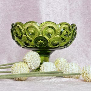 May include: A green glass bowl with a pedestal base and a textured surface. The bowl is decorated with a repeating pattern of circles. The bowl is surrounded by white flowers.