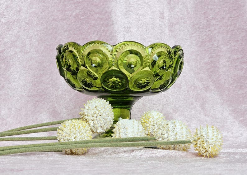 May include: A green glass bowl with a pedestal base and a textured surface. The bowl is decorated with a repeating pattern of circles. The bowl is surrounded by white flowers.