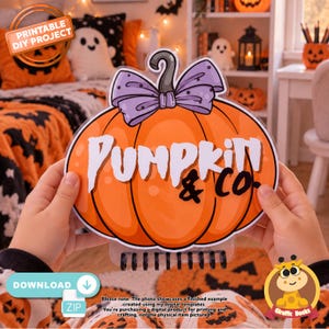 以下が含まれることがあります： 上部に紫色のリボン、中央に白と黒で「PUMPKIN & CO.」と書かれたカボチャ型のサイン。下部には黒いスパイラル綴じがあり、両手で持たれた状態で、印刷可能なDIYプロジェクトのデザインを示しています。