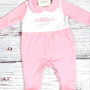 May include: A pink baby romper with long sleeves and attached feet. The romper has a white bib-like panel with the name "Millie" embroidered in pink script, flanked by two hearts. A light pink Peter Pan collar completes the design.