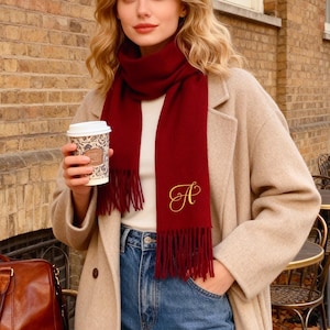 May include: A woman wearing a long beige coat, blue jeans, and a burgundy scarf with an embroidered initial. She holds a coffee cup. A brown leather duffel bag and beige knit gloves sit on a black table. The background features a brick building and outdoor seating.