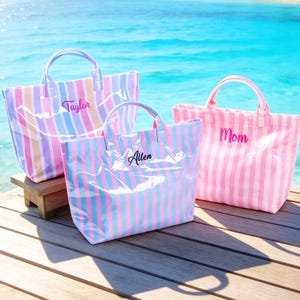 May include: Three striped tote bags with handles, each with a name embroidered on the front. The bags are in pastel colors of pink, blue, and yellow. The background shows a tropical beach scene with turquoise water and palm trees.