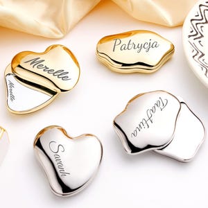 May include: A collection of personalized compact mirrors in gold and silver tones. The mirrors are in various shapes, including heart and organic forms, with engraved names. One mirror is open, revealing a reflective surface.