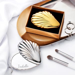 Puede incluir: Un espejo compacto dorado en forma de concha en una caja marr&oacute;n, y un espejo compacto plateado en forma de concha con el nombre "Isabella" grabado. Tambi&eacute;n se ven dos brochas de maquillaje y un par de anillos.
