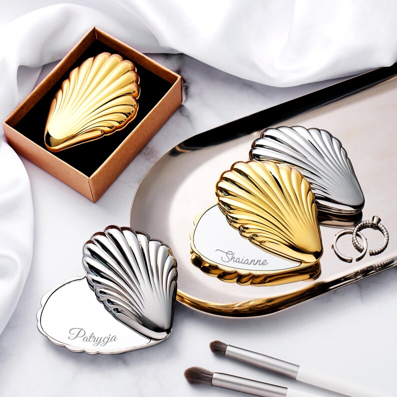 Puede incluir: Espejos compactos en forma de concha en tonos dorados y plateados. Un compacto dorado est&aacute; en una caja marr&oacute;n. Tambi&eacute;n hay dos anillos plateados y dos brochas de maquillaje. Los compactos tienen nombres personalizados.