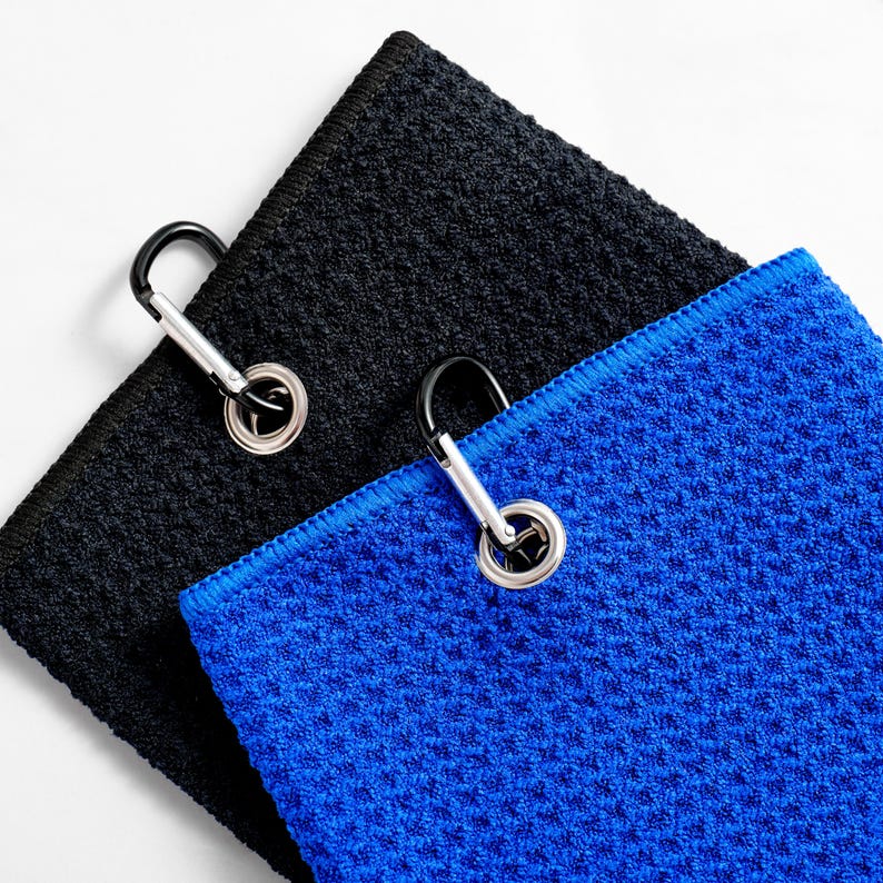 May include: Two golf towels, one black and one blue, are shown. Each towel has a textured surface and a silver carabiner clip attached. The blue towel has a blue border. The towels are angled against a white background.
