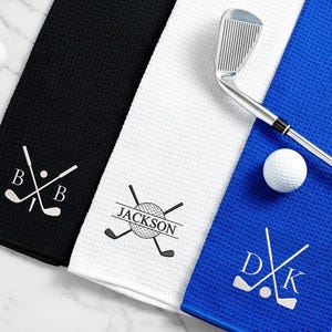 May include: Three golf towels in black, white, and blue, with personalized embroidered designs. The black towel has "B B" initials, the white towel says "Jackson", and the blue towel has "D K" initials. Golf balls and a golf club are also present.