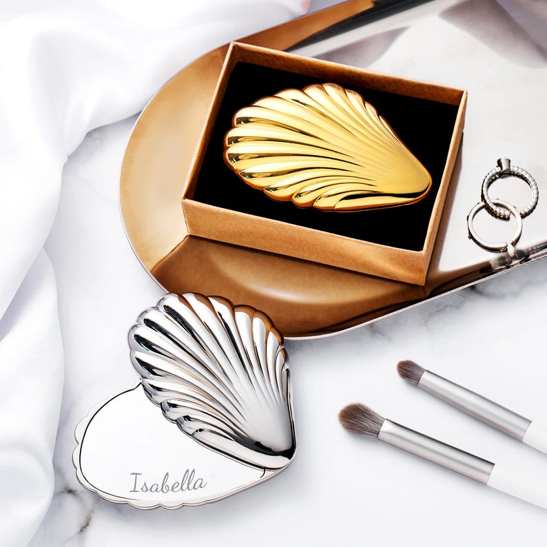 Puede incluir: Un espejo compacto dorado en forma de concha en una caja marr&oacute;n, y un espejo compacto plateado en forma de concha con el nombre "Isabella" grabado. Tambi&eacute;n se ven dos brochas de maquillaje y un par de anillos.