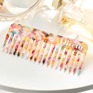 May include: A colorful comb with the name "Olivia" in gold lettering. The comb features a vibrant pattern of pink, yellow, green, and blue hues. The comb is on a white surface, with a mirror and perfume bottle in the background.