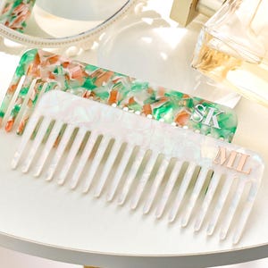 May include: Two hair combs. One is white with a pearlescent finish, the other is green, orange, and white. The white comb has the initials "MIL" in gold. The green comb has the initials "SK" in silver.