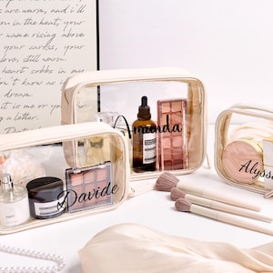 Op de afbeelding: Drie transparante cosmetische tassen met beige randen, elk met make-upproducten. De tassen zijn gelabeld met "Amanda", "Davide" en "Alyssa". Make-up borstels zijn vooraan geplaatst. Op de achtergrond staat een handgeschreven gedicht.