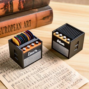 May include: Two miniature black and orange guitar amplifier-shaped boxes with the names "Laura" and "Oliver" on the front. One holds colorful guitar picks. The boxes sit on sheet music.