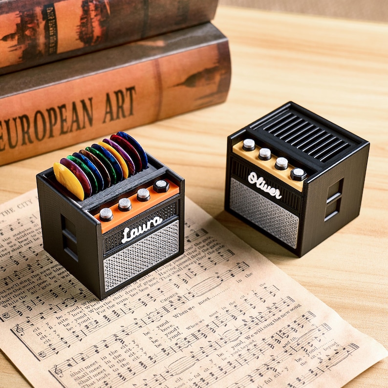 May include: Two miniature black and orange guitar amplifier-shaped boxes with the names "Laura" and "Oliver" on the front. One holds colorful guitar picks. The boxes sit on sheet music.