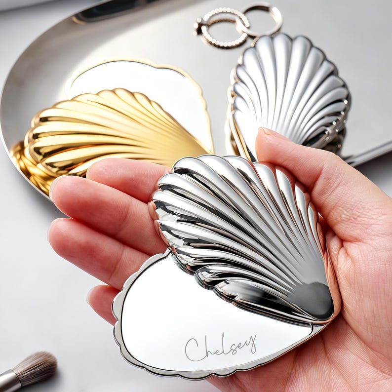 Puede incluir: Un espejo compacto plateado en forma de coraz&oacute;n y con forma de concha, abierto para revelar una superficie de espejo con el nombre "Chelsey" grabado. Un compacto dorado en forma de concha y un anillo plateado est&aacute;n en el fondo.
