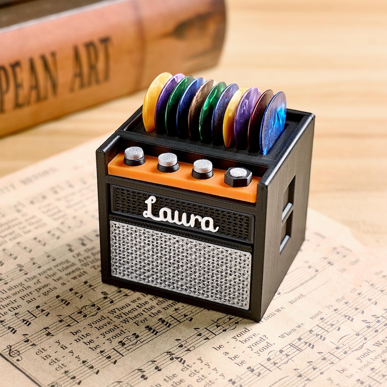 May include: A black and orange miniature guitar amplifier with the name "Laura" on the front. The amplifier holds a collection of colorful guitar picks. The background includes sheet music and a wooden object.
