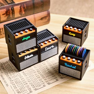 May include: Small black and orange guitar amplifier-style boxes, each with a name such as "Angie" or "Lucy". Some boxes hold colourful guitar picks. The boxes are displayed on a sheet of musical notation.