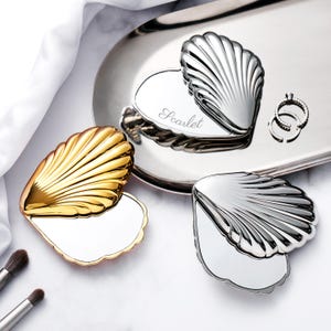 May include: Three seashell-shaped compact mirrors in gold and silver tones, arranged on a silver tray. One mirror is open, revealing a reflective surface. The word "Scarlet" is engraved on one of the compacts. Two rings are also on the tray.