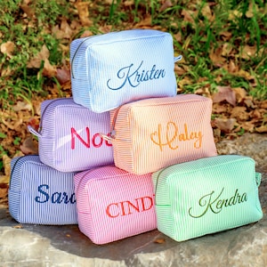 May include: A stack of six seersucker cosmetic bags in various colors: light blue, purple, orange, pink, and green. Each bag has a different name embroidered on it in a matching color. The bags have a rectangular shape with a zipper closure.