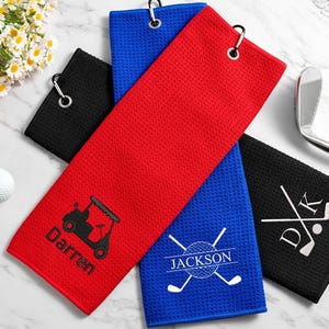 May include: Golf towels in red, blue, and black with custom embroidery. The red towel shows a golf cart and the name "Darren." The blue towel has crossed golf clubs and the name "Jackson." The black towel displays initials and crossed clubs.