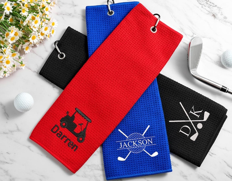 May include: Golf towels in red, blue, and black with custom embroidery. The red towel shows a golf cart and the name "Darren." The blue towel has crossed golf clubs and the name "Jackson." The black towel displays initials and crossed clubs.