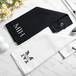 May include: Two golf towels, one black and one white, are shown. The black towel features the initials "MIH" in white, and the white towel displays the name "MEHTA" in black. Two golf balls and a golf club are also present.