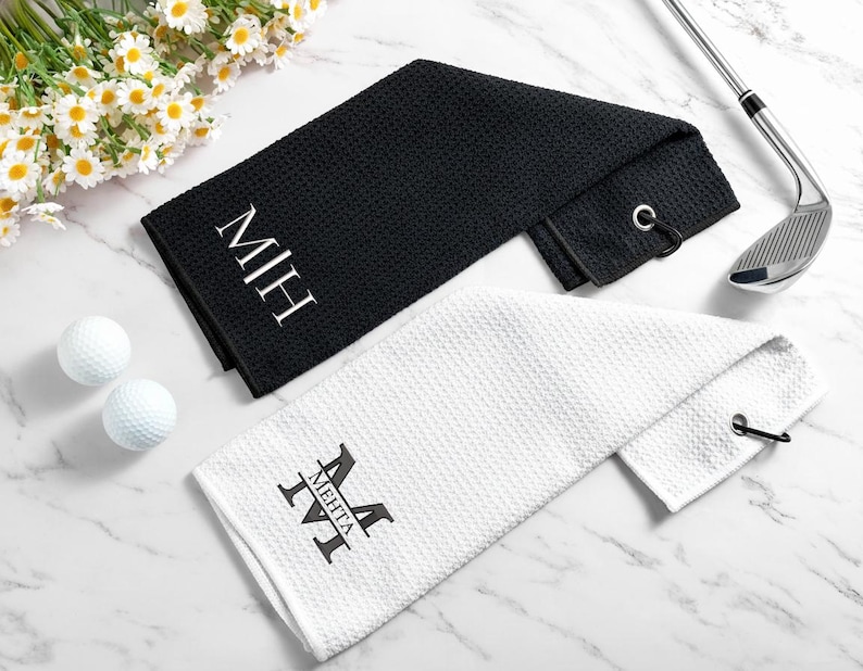 May include: Two golf towels, one black and one white, are shown. The black towel features the initials "MIH" in white, and the white towel displays the name "MEHTA" in black. Two golf balls and a golf club are also present.