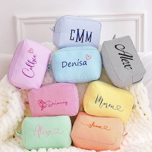 Può includere: Una collezione di trousse personalizzate in vari colori, tra cui rosa, blu, giallo e arancione. Ogni borsa presenta un nome o delle iniziali ricamate in corsivo. Le borse hanno un motivo a righe.