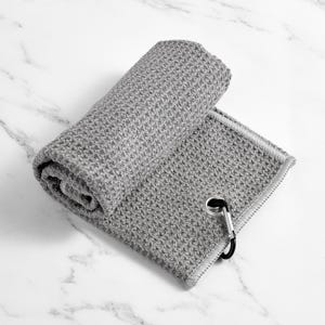 May include: A rolled-up grey golf towel with a textured waffle weave. The towel has a silver-coloured grommet and a black carabiner clip for easy attachment to a golf bag. The towel is folded over on a white marble surface.